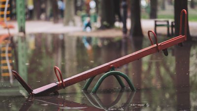 A bright red seesaw tilted awkwardly in the flooded area of a park after a heavy downpour