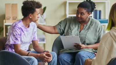 African young psychologist talking to teenage boy during therapy session at classroom