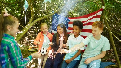 Multi-ethnic group of kids lighting sparklers while hiding under branches of big tree