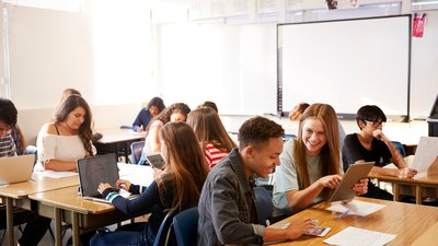 Wide Angle View Of High School Students Sitting At Desks In Classroom Using Laptops